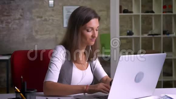 有吸引力的积极女性办公室工作人员在耳机里听音乐在工作场所的笔记本电脑上打字视频的预览图