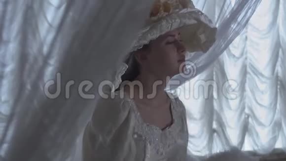 穿着白色老式球裙和白色帽子站在透明窗帘和视频的预览图