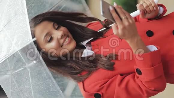 垂直视频美丽的年轻商务女性在雨天的街道上使用智能手机微笑着抱着视频的预览图