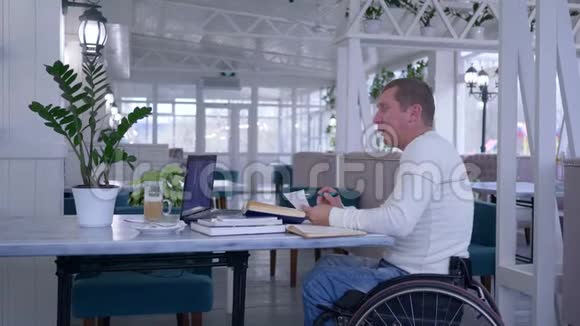 在线教育残疾人坐在轮椅上用手提电脑说话坐在桌子上拿着书和一杯咖啡视频的预览图