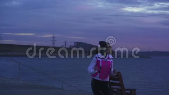 下面的动作一个穿着运动帽和夹克的年轻女孩在黎明前在河堤上晨跑视频的预览图