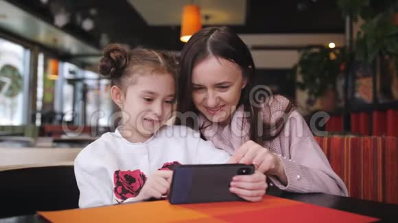 漂亮的白种人母亲和她的女儿在咖啡馆里使用智能手机一个先进的小女孩展示并解释视频的预览图