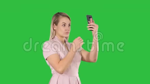 一个金发女人在绿色屏幕上的智能手机前梳妆打扮色度键视频的预览图