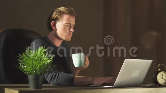 年轻的金发商人在现代办公室的笔记本上工作喝着咖啡视频的预览图
