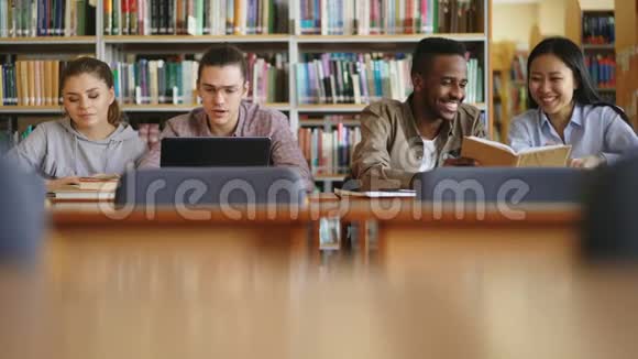 多民族学生坐在图书馆桌上放着书和笔记本电脑一起准备考试视频的预览图