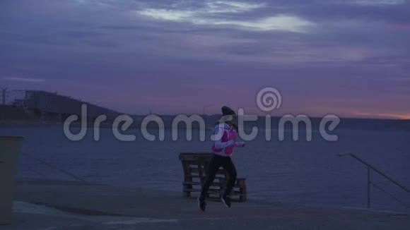 下面的动作一个穿着运动帽和夹克的年轻女孩在黎明前在河堤上晨跑视频的预览图