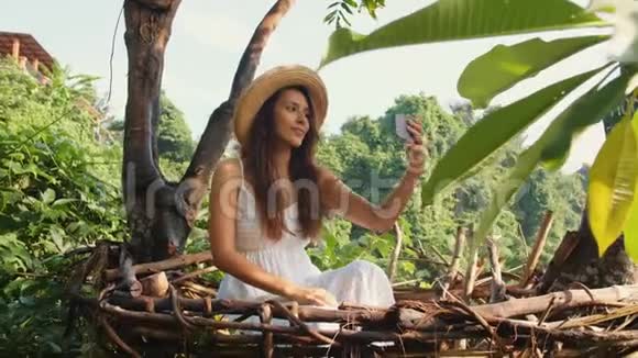 年轻的微笑混合种族旅游女孩穿着白色连衣裙制作自拍照片使用手机坐在装饰草巢视频的预览图
