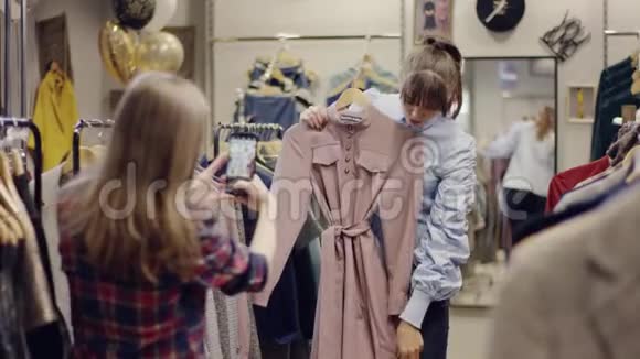 年轻可爱的女人在智能手机上拍照她的女朋友在服装店试穿衣服视频的预览图