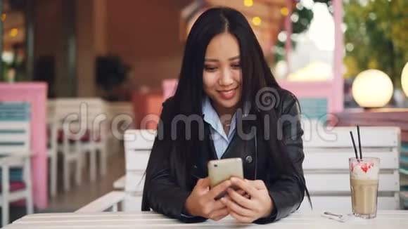 微笑的亚洲女孩学生正在户外咖啡馆休息使用智能手机触摸屏幕和观看社会媒体视频的预览图