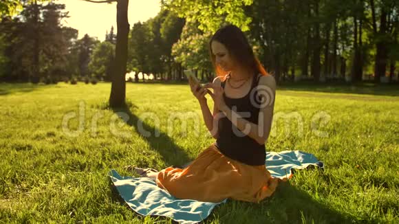 红头发的女孩享受阳光明媚的一天使用手机坐在绿色夏季公园的长凳上视频的预览图