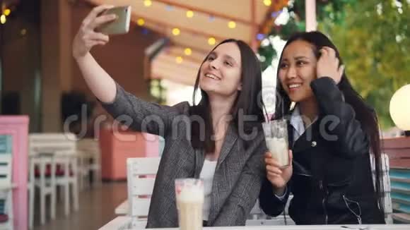 迷人的女孩正和她的亚洲朋友一起自拍她坐在露天咖啡馆里拿着智能手机和饮料视频的预览图