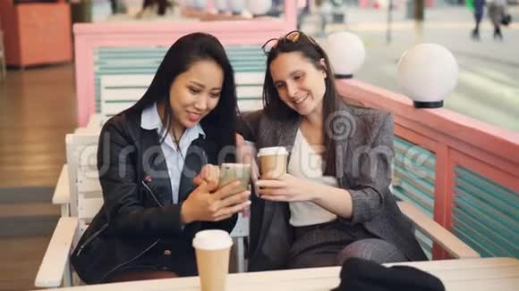 漂亮的年轻女士朋友们正在使用现代智能手机坐在咖啡馆的桌子上带走咖啡快乐的女孩视频的预览图
