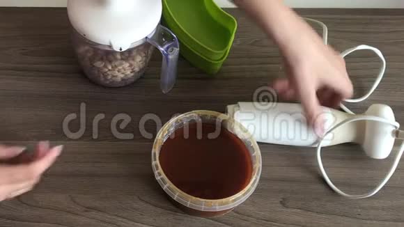 一个女人关上搅拌器碗的盖子研磨花生和种子来制作卤汁旁边的容器是蜂蜜加入视频的预览图
