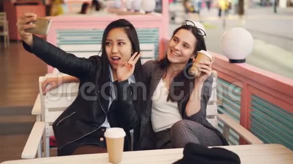 无忧无虑的女孩正在自拍带着外卖的饮料坐在咖啡馆里一起使用智能手机年轻女性视频的预览图