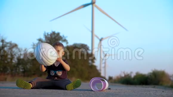 一个孩子玩建筑头盔坐在道路上背景上有风力发电机慢动作视频的预览图