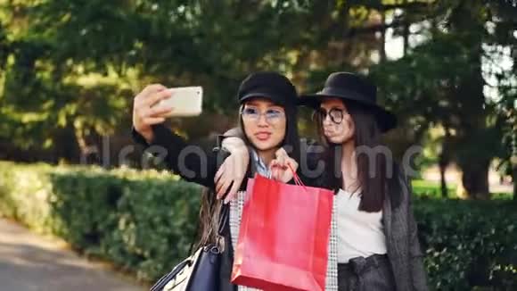 漂亮的亚洲女孩正和她的高加索朋友在购物一天后拿着袋子用智能手机自拍视频的预览图