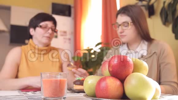 妈妈和带眼镜的小女儿在厨房喝茶或咖啡吃蛋糕说话笑在前景中视频的预览图