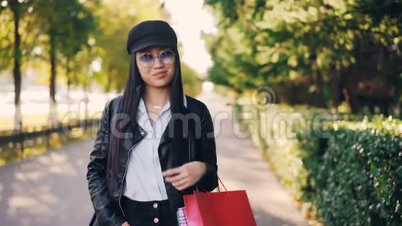 戴着皮夹克和帽子看着相机微笑着拿着购物袋的可爱亚洲女孩的肖像视频的预览图