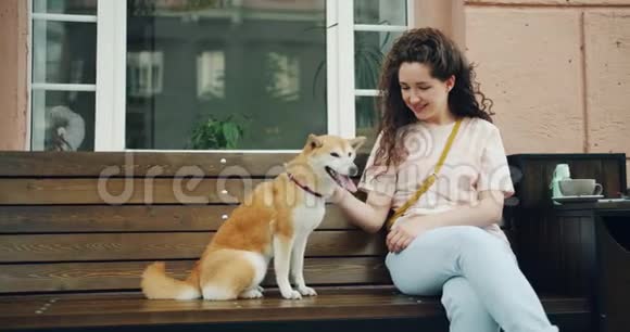 快乐的年轻女人在咖啡馆里抚摸着可爱的石巴狗小狗坐在户外的长凳上视频的预览图