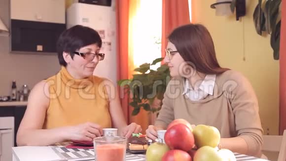 妈妈和带眼镜的小女儿在厨房喝茶或咖啡吃蛋糕说话笑在前景中视频的预览图