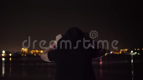 夜晚一个穿着黑色引擎盖的男人正在跳舞从后面看在水面上发光的城市的灯光下视频的预览图