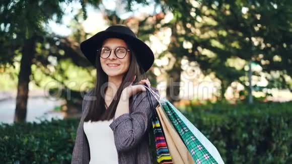 肖像好看的女孩戴着帽子和眼镜拿着购物袋看着相机微笑着优雅优雅视频的预览图