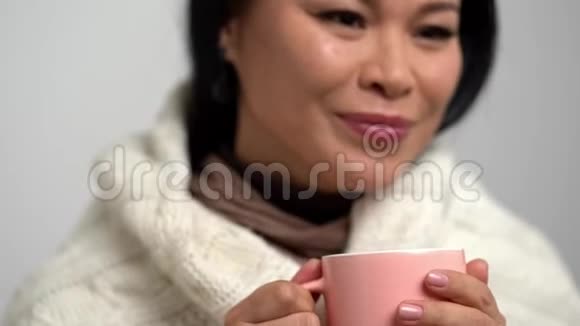 女人带着甜蜜的微笑包在一个温暖的针织围巾喝茶从粉红杯视频的预览图