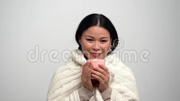 亚洲女人带着甜蜜的微笑裹着温暖的针织围巾喝着粉红杯里的茶视频的预览图