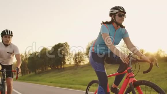 两个骑自行车的人一男一女在公路上骑自行车在日落时分他们穿着头盔和运动服慢动作视频的预览图
