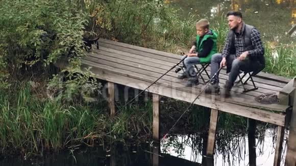 英俊的男人和他可爱的儿子正在池塘里钓鱼从木坞坐在椅子上拿着棍子说话爱视频的预览图