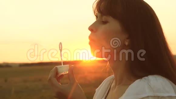 美丽的女孩在春天夏天和微笑的夕阳下在公园里吹着美丽的肥皂泡慢动作年轻青年视频的预览图