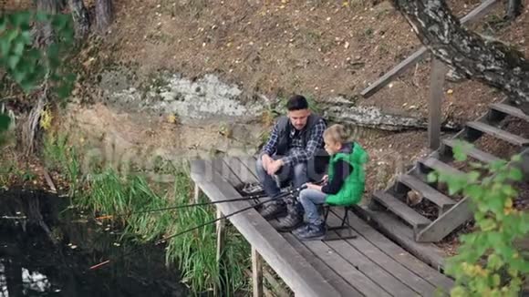 小男孩和爸爸在秋日从木墩上钓鱼小男孩正拿着棍子坐在椅子上视频的预览图