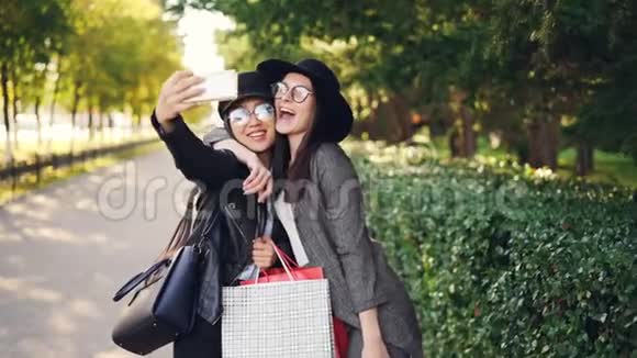 兴奋的女性朋友正在使用智能手机自拍并在阳光明媚的秋日拿着购物袋购物视频的预览图