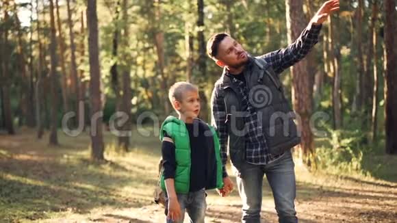 爱的爸爸在阳光明媚的秋日和儿子谈论大自然时站在森林里做着手势微笑着视频的预览图
