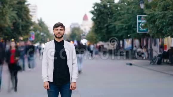 一个穿着牛仔裤和白夹克的自信男人独自站在城市的街道上一边看着镜头时间流逝视频的预览图