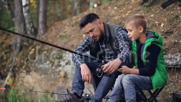帅哥爱爸爸正在教儿子一起钓鱼坐在河岸上男孩正拿着鱼竿移动视频的预览图