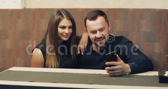 一个留着胡子的男人和一个漂亮的黑发女人坐在酒吧里看电话视频的预览图