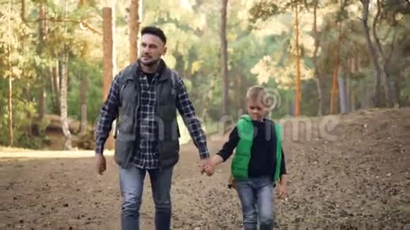 可爱的男孩正在森林里散步他慈爱的父亲牵着他的手蓄着胡子的年轻人正在说话和做手势视频的预览图