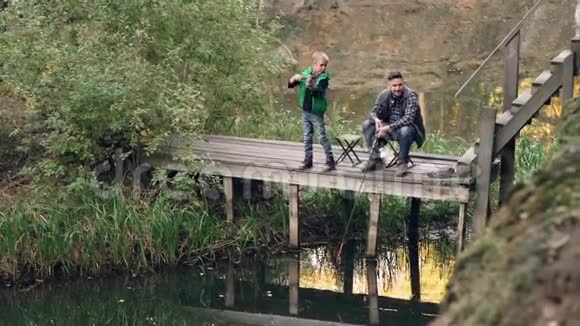 严肃的孩子站在木墩和钓鱼竿上而他慈爱的父亲则坐在椅子上抓鱼视频的预览图