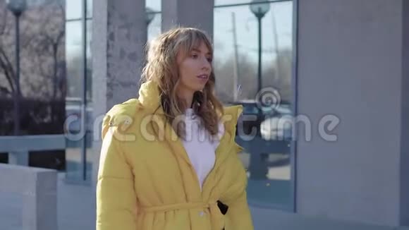 秋天年轻迷人的金发女郎穿着时尚的黄色外套在城市里散步路过一座建筑视频的预览图