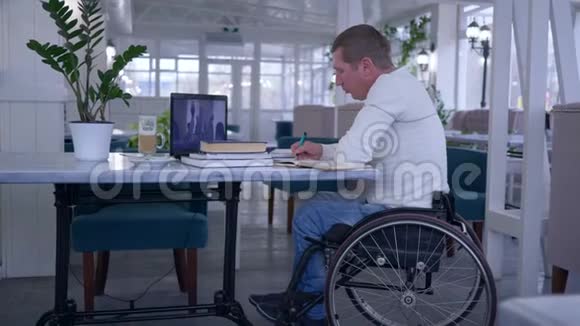 在线培训残疾学生在轮椅上做笔记在远程教育期间坐在桌子上视频的预览图