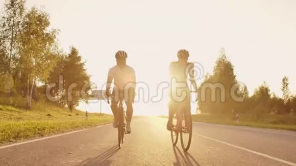 两个骑自行车的人一男一女在公路上骑自行车在日落时分他们穿着头盔和运动服慢动作视频的预览图