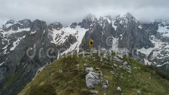一个徒步旅行者站在山顶上看着白雪皑皑的山脉举起双手做胜利的手势视频的预览图