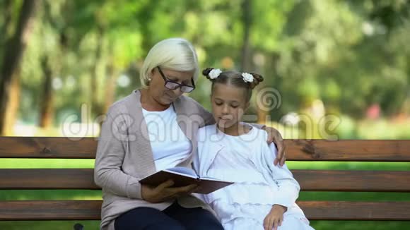 照看奶奶给坐在公园长椅上的可爱孙女读童话视频的预览图
