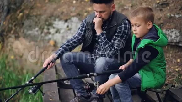 严肃的小男孩正和他的父亲长着胡须的黑发坐在一起拿着钓鱼竿钓鱼共同视频的预览图