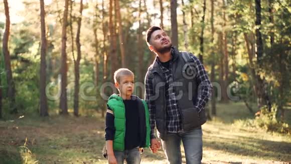 好奇的孩子和他的父亲留着胡子的家伙在阳光明媚的白天站在森林里看着大自然视频的预览图