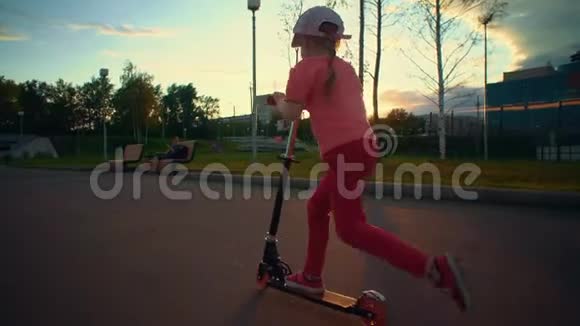 小可爱的金发女孩穿着粉红色的帽子在公共公园骑滑板车视频的预览图
