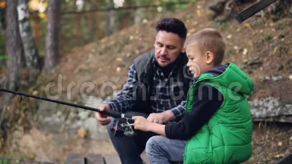 可爱的小男孩正和爸爸在森林里的池塘里钓鱼爸爸正在跟他解释怎么做视频的预览图