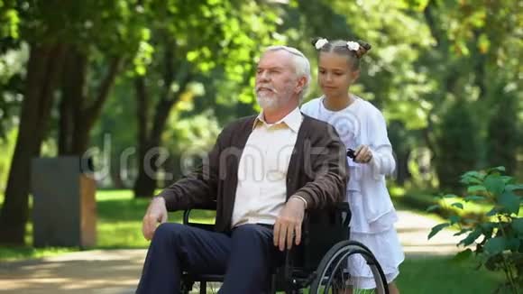 可爱的女孩拥抱坐在轮椅上的祖父在公园里散步消遣视频的预览图