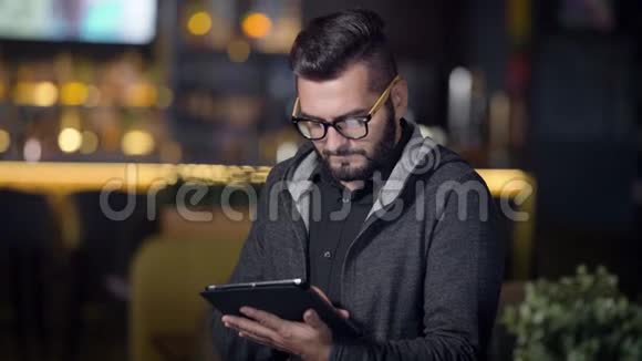 穿着时髦发型戴着眼镜的成年人晚上坐在酒吧里用平板电脑上网视频的预览图